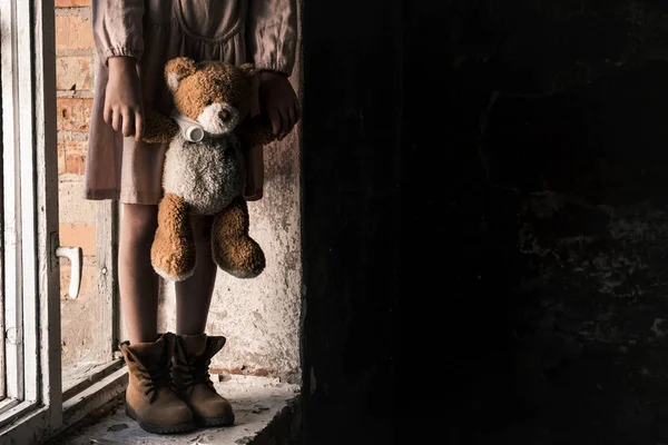 Cropped view of child holding dirty teddy bear while standing on windowsill, post apocalyptic concept — Stock Photo