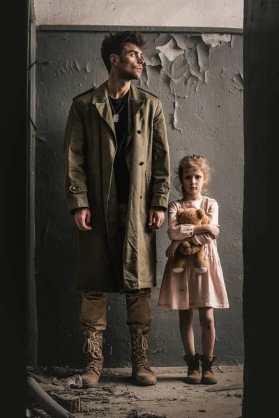 Handsome man standing near kid with teddy bear near weathered wall, post apocalyptic concept — Stock Photo