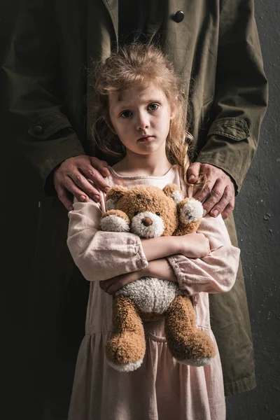 Обрізаний вид людини, що стоїть біля дитини з плюшевим ведмедем, пост-апокаліптична концепція — стокове фото