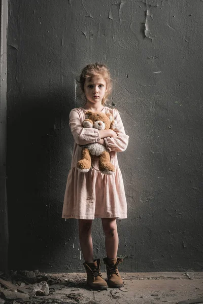 Upset kid looking at camera while standing with soft toy near wall, post apocalyptic concept — Stock Photo