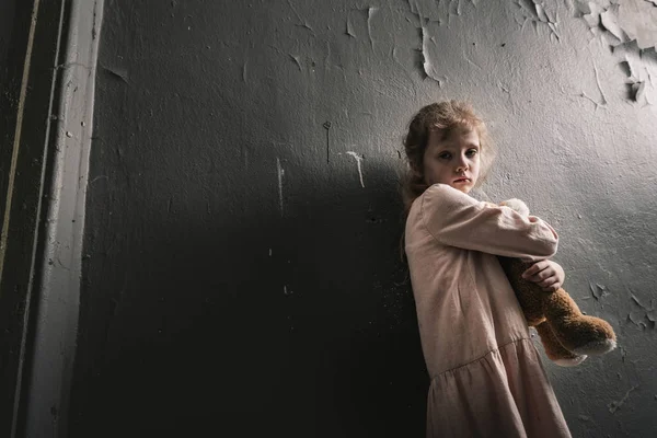 Расстроенный ребенок смотрит в камеру, стоя с мягкой игрушкой, пост апокалиптической концепции — стоковое фото