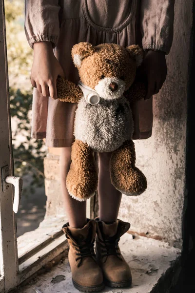 Vista recortada de niño sosteniendo oso de peluche sucio cerca de la ventana, concepto post apocalíptico - foto de stock