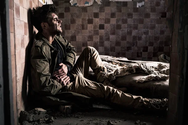 Side view of man with dirty face having stomach ache while sitting on floor, post apocalyptic concept — Stock Photo