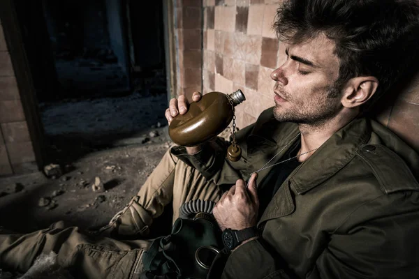 Hombre guapo sosteniendo frasco mientras está sentado en un edificio abandonado, concepto post apocalíptico - foto de stock