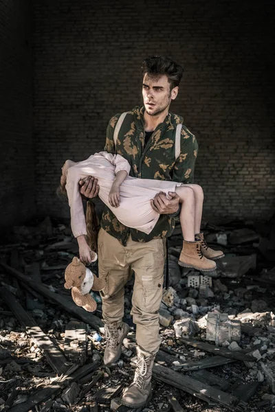Hombre molesto sosteniendo en brazos niño con osito de peluche cerca de edificio abandonado, concepto post apocalíptico - foto de stock
