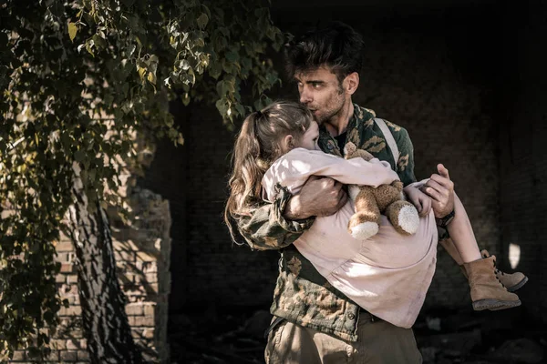 Hombre guapo sosteniendo en brazos y besando niño con osito de peluche, concepto post apocalíptico - foto de stock