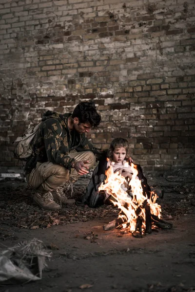 Селективное внимание мужчины в куртке на ребенка, сидящего у костра, постапокалиптическая концепция — стоковое фото