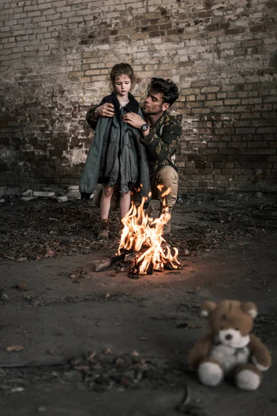 Enfoque selectivo del hombre que usa chaqueta en el niño cerca de la hoguera, concepto post apocalíptico - foto de stock