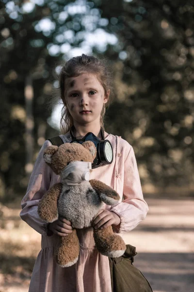 Грязный ребенок держит грязную мягкую игрушку рядом с деревьями в Чернобыле, пост-апокалиптическая концепция — стоковое фото