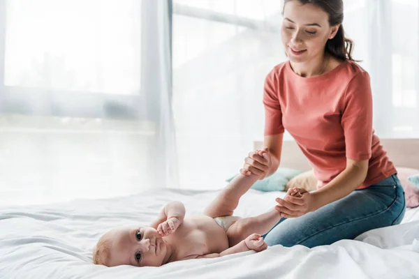 Glückliche Mutter berührt Beine des Babys, das auf dem Bett liegt — Stockfoto