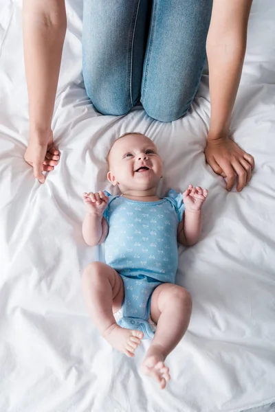 Обрізаний вид матері в джинсах, що сидить біля щасливого немовляти в блакитному дитячому боді — стокове фото