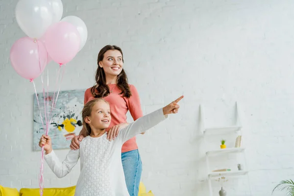 Niño feliz señalando con el dedo mientras sostiene globos rosados cerca de niñera - foto de stock