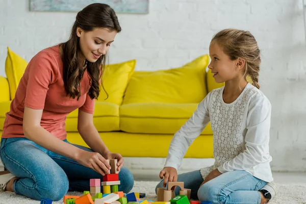 Happy kid looking at babysitter near toy blocks on carpet — Stock Photo