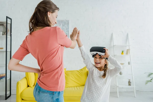 Enfant gai dans le casque de réalité virtuelle donnant haute cinq à baby-sitter à la maison — Photo de stock