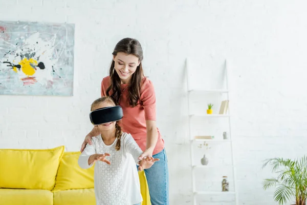 Niñera feliz de pie con un lindo niño en auriculares de realidad virtual - foto de stock