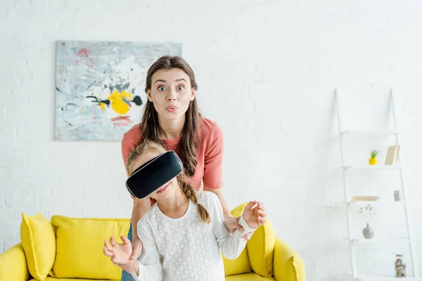 Niñera mirando a la cámara cerca de niño en auriculares de realidad virtual - foto de stock