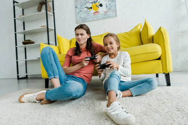 KYIV, UCRANIA - 4 de septiembre de 2019: niñera emocional y niño feliz sentado en la alfombra y jugando videojuegos en la sala de estar - foto de stock