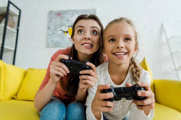 KYIV, UCRANIA - 4 de septiembre de 2019: niñera mordiendo labios cerca de un niño feliz mientras juega videojuegos en la sala de estar - foto de stock