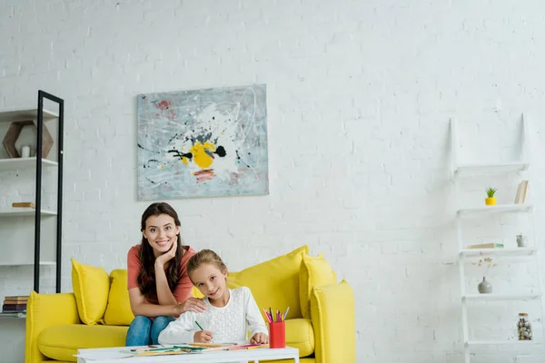 Happy babysitter sitting on yellow sofa near kid drawing in living room — Stock Photo