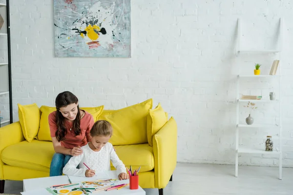 Fröhliche Babysitterin sitzt auf gelbem Sofa neben Kind Zeichnung im Wohnzimmer — Stockfoto