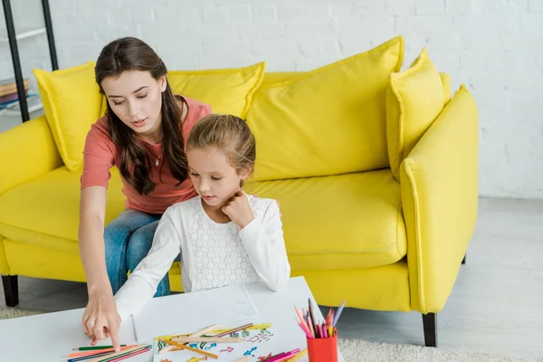 Привлекательная няня сидит на желтом диване рядом с ребенком и цветные карандаши в гостиной — стоковое фото