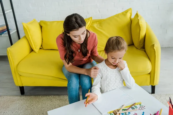 Attraente babysitter seduta sul divano giallo vicino al bambino guardando disegno disegnato in soggiorno — Foto stock