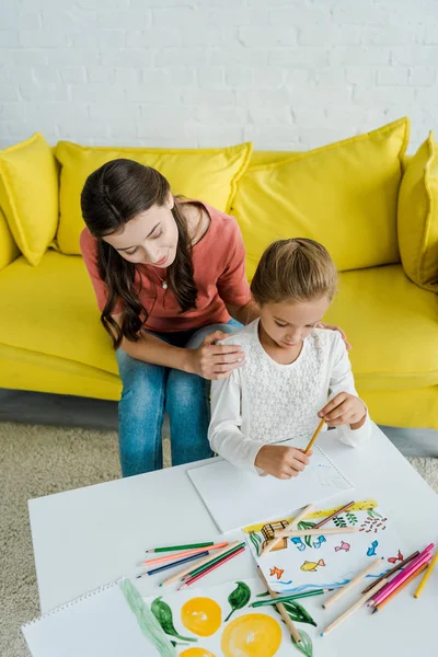 Beautiful babysitter sitting on yellow sofa near kid looking at drawn pictures in living room — Stock Photo