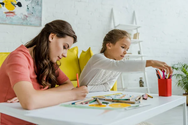 Селективный фокус привлекательной няни рисования рядом с симпатичным ребенком в гостиной — стоковое фото
