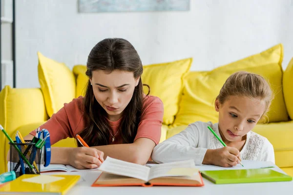 Selective focus of babysitter near kid sticking out tongue while studying in living room — Stock Photo