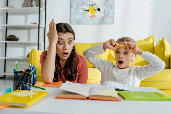 Selective focus of shocked kid looking at book near surprised babysitter in living room — Stock Photo