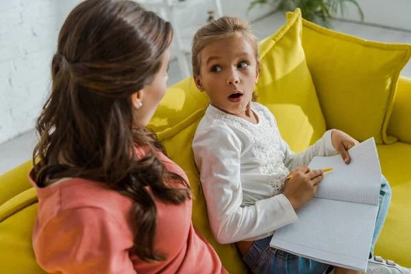 Surprised kid holding notebook and looking at babysitter in living room — Stock Photo