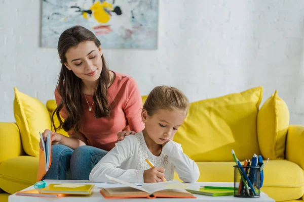 Selective focus of happy babysitter sitting on sofa and looking at cute kid studying at home — Stock Photo