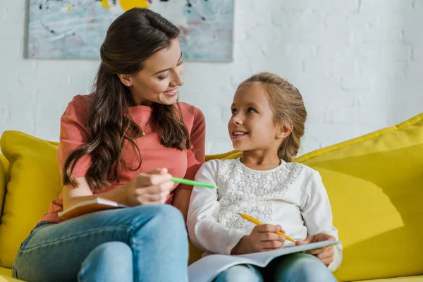 Selektiver Fokus des glücklichen Kindes mit Notizbuch und Blick auf fröhlichen Babysitter mit Bleistift im Wohnzimmer — Stockfoto