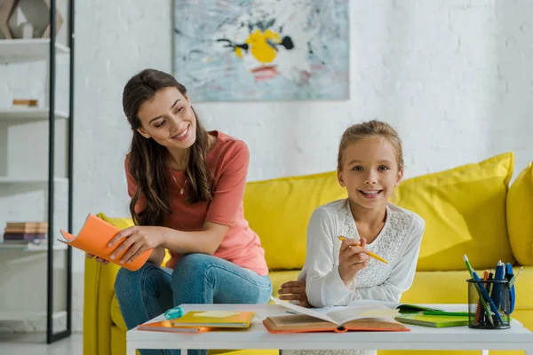 Selective focus of happy babysitter holding notebook while sitting on sofa and looking at cute kid studying at home — Stock Photo