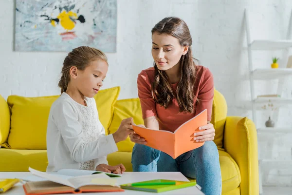 Selektiver Fokus eines attraktiven Babysitters, der ein Notizbuch in der Nähe eines niedlichen Kindes hält, das zu Hause studiert — Stockfoto
