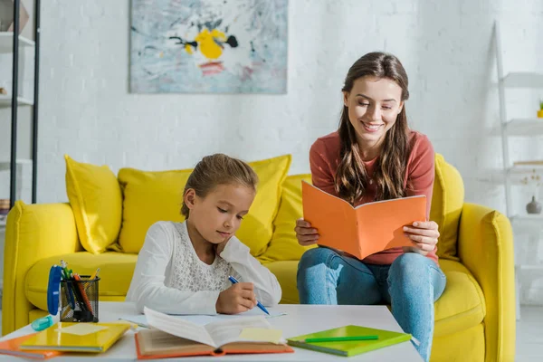 Foyer sélectif de baby-sitter attrayant tenant carnet près de mignon enfant étudiant à la maison — Photo de stock