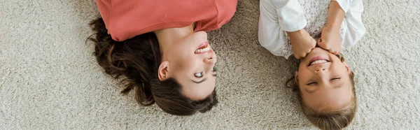 Panoramic shot of happy babysitter looking at kid lying on carpet — Stock Photo
