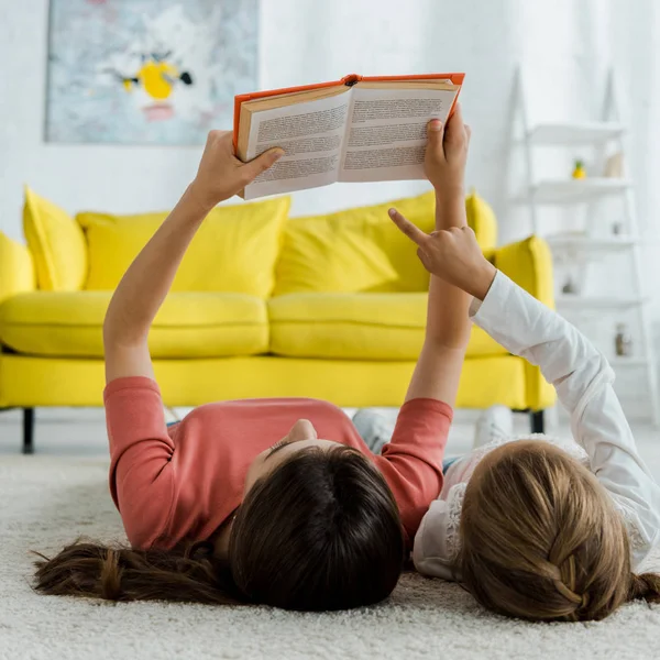 Kind zeigt mit Finger auf Buch, während es mit Babysitter auf Teppich liegt — Stockfoto