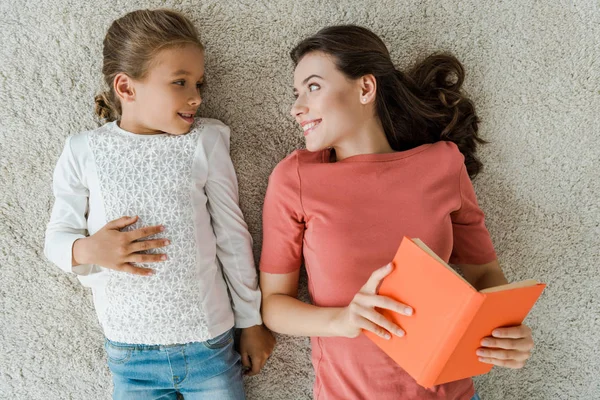 Top view of cheerful babysitter holding book while looking at kid lying on carpet — Stock Photo