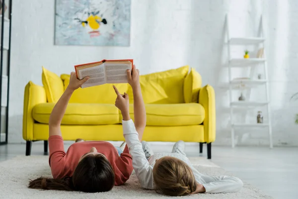 Kind zeigt mit Finger auf Buch, während es mit Babysitter im Wohnzimmer auf Teppich liegt — Stockfoto