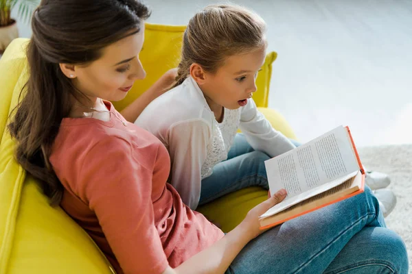 Selektiver Fokus des überraschten Kindes auf dem Sofa sitzend mit glücklichem Babysitter, der Buch liest — Stockfoto