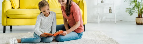 Panoramic shot of kid reading book while sitting on carpet with babysitter — Stock Photo