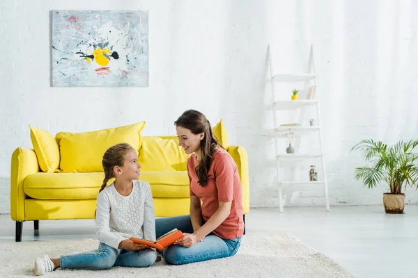 Happy kid holding book while sitting on carpet with babysitter — Stock Photo