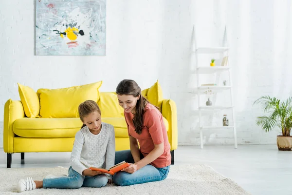 Feliz niño leyendo libro mientras está sentado en la alfombra con niñera - foto de stock