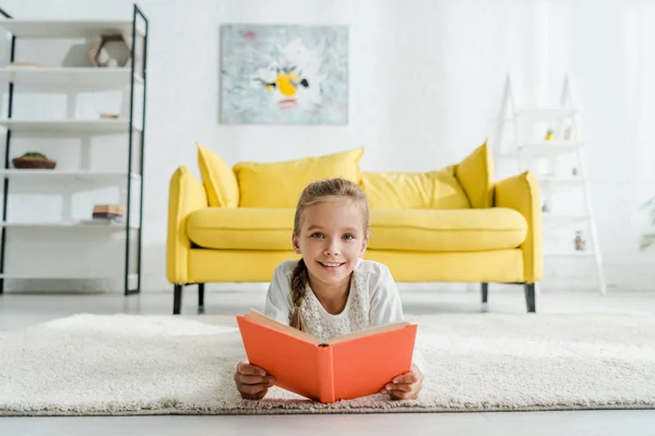 Happy kid holding book white lying on carpet near yellow sofa — Stock Photo