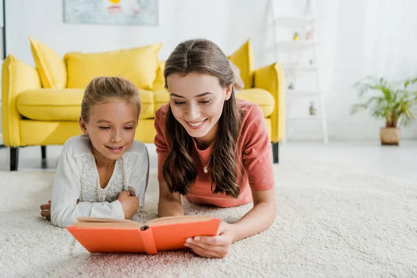 Happy babysitter lying on carpet with cheerful kid while reading book — Stock Photo