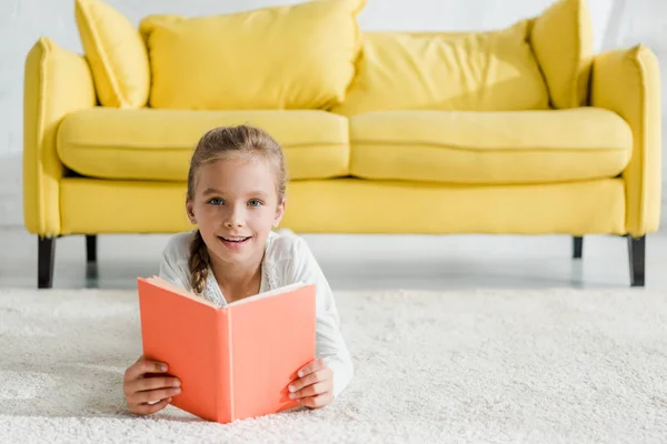 Heureux enfant tenant livre blanc couché sur le tapis près du canapé jaune — Photo de stock
