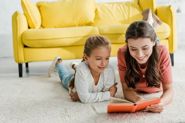 Happy babysitter lying on carpet with smiling kid while reading book — Stock Photo