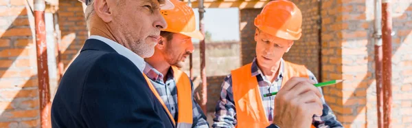 Панорамный снимок бородатого взрослого мужчины рядом со строителями в шлемах — стоковое фото