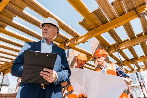 Low angle view of bearded mature businessman with clipboard near builders — Stock Photo